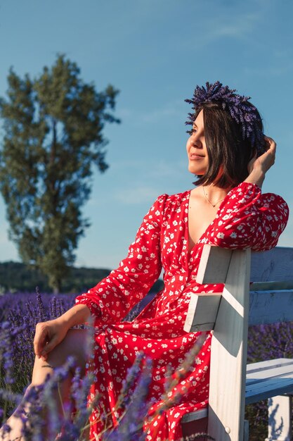 A young woman in a wreath on her head sits on a white bench in a lavender field