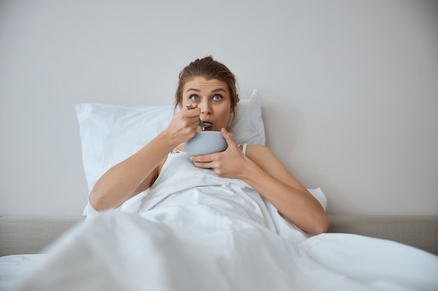 Young woman wrinkling her forehead while looking upwards and eating soup