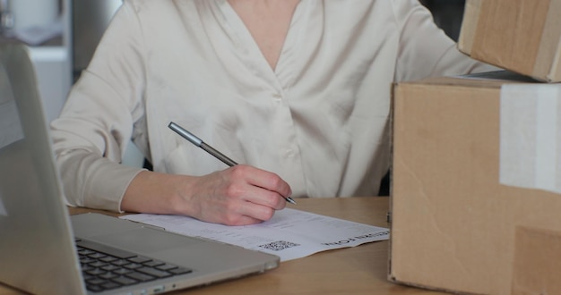 若い女性が返品注文を書き、自宅でノートパソコンを持ってテーブルに座っている 紙に書いて、段ボール箱を返品したいフォームに記入している女性顧客のクローズアップビデオ