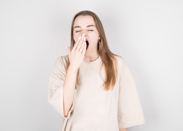 Young woman yawns from fatigue near the gray wall