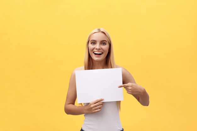 Photo young woman over yellow background holding blank paper sheet with surprise expression pointing finger copy space.