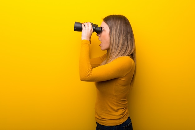 Young woman on yellow background and looking in the distance with binoculars