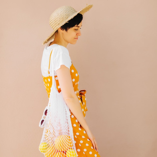 Young woman in yellow clothes and a straw hat with a mesh bag of fruit on light beige background