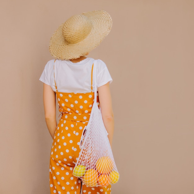 Young woman in yellow clothes and a straw hat with a mesh bag of fruit on light beige background