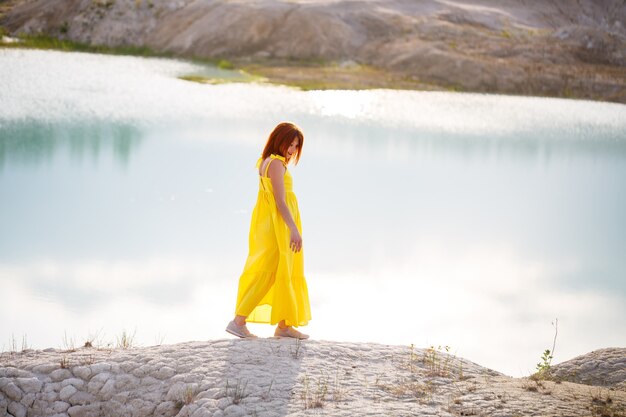 Young woman in a yellow dress near the lake with azure water and green trees.