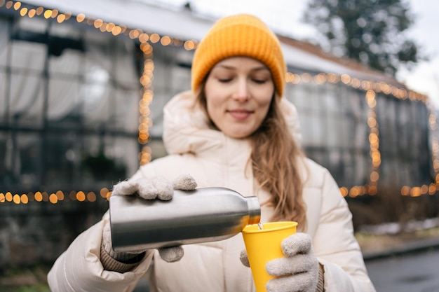 A young woman in a yellow hat pours tea from a metal thermos into a yellow glass on a background of garlands The concept of spending time outdoors and enjoying winter