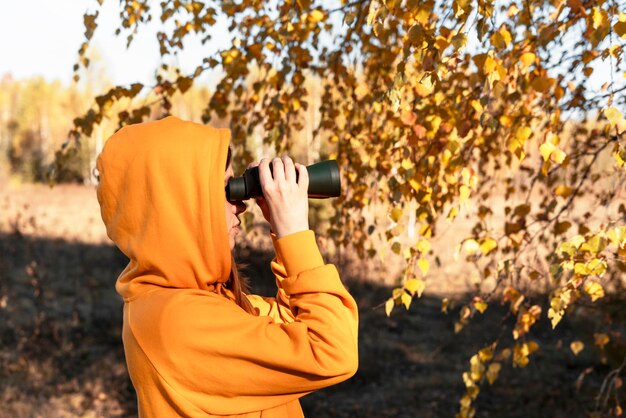 Photo young woman in yellow hood with binoculars watching birds in the autumn forest birdwatching