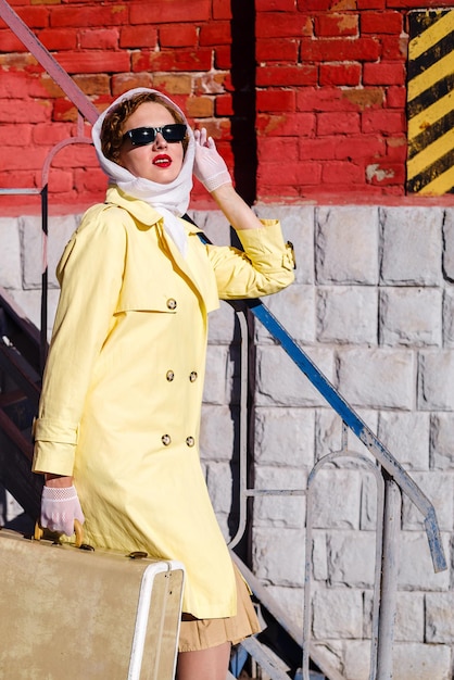 Foto giovane donna in impermeabile giallo e occhiali scuri con la valigia in mano contro un muro di mattoni