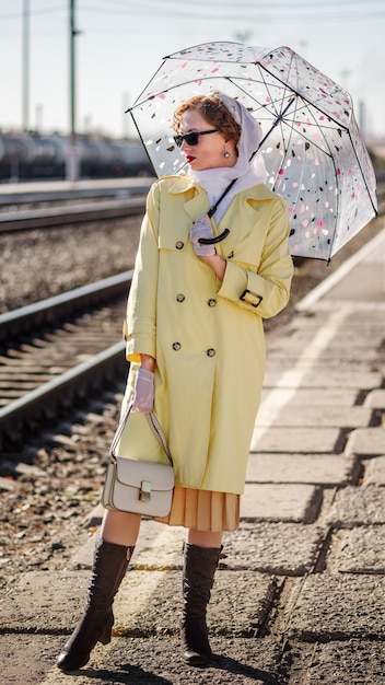 Foto giovane donna in impermeabile giallo e occhiali scuri con ombrello e borsetta sulla stazione ferroviaria