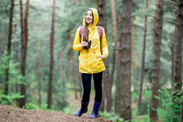 Young woman in yellow raincoat hiking with binoculars and backpack in the green pine forest. Bird watching in the forest