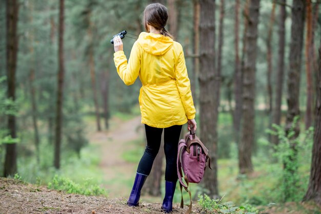 Young woman in yellow raincoat standing with backpack in the green pine forest