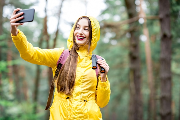 Young woman in yellow raincoat taking selfie portrait while hiking with binoculars and backpack in the green forest