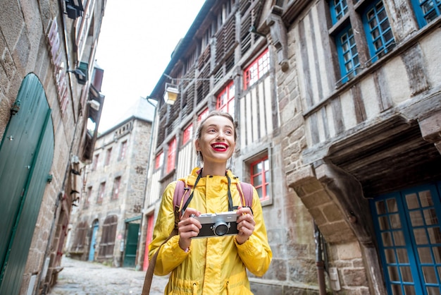 Молодая женщина в желтом плаще гуляет с рюкзаком и фотоаппаратом в деревне Динан в регионе Бретань во Франции