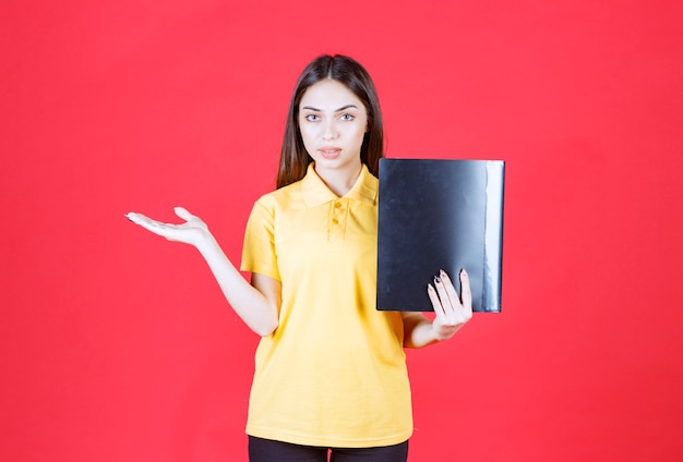 Young woman in yellow shirt holding a black folder, pointing and calling her colleague