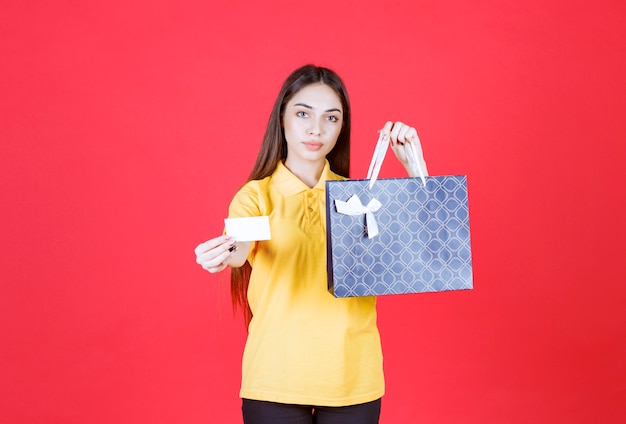 Young woman in yellow shirt holding a blue shopping bag and presenting her business card