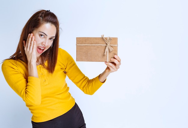 Молодая женщина в желтой рубашке держит картонную подарочную коробку и выглядит удивленной