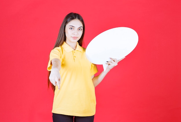 Young woman in yellow shirt holding an ovale info board and inviting her colleague next to her