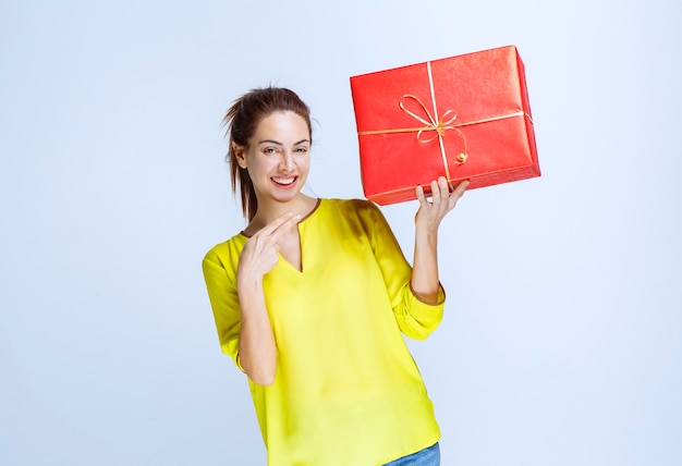 Young woman in yellow shirt holding a red gift box and pointing at it