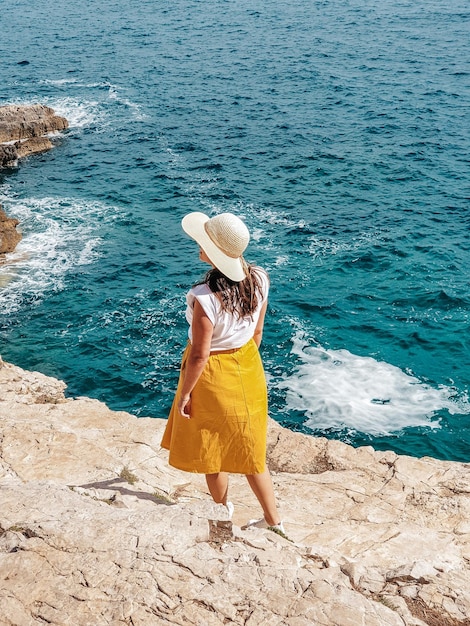 Young woman in yellow skirt standing on cliff above sea