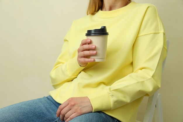 Young woman in yellow sweatshirt sitting with paper cup against\
beige surface