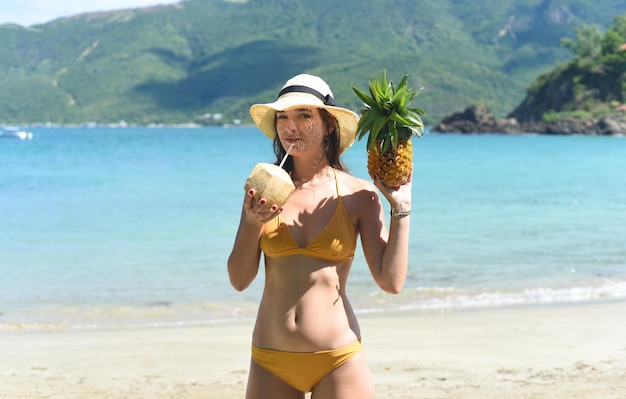 Young woman in yellow swimsuit drinking coconut milk and holding pineapple on sandy beach