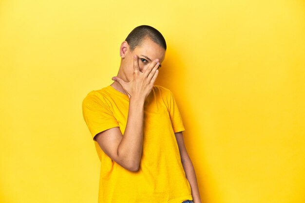 Photo young woman in yellow tee yellow studio backdrop blink at the camera through fingers