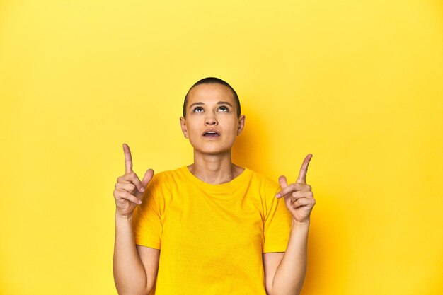 口を開いて逆さまを指す黄色の t シャツの黄色のスタジオ背景の若い女性