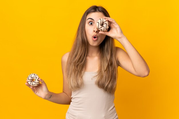 Young woman over yellow wall holding a donut in an eye