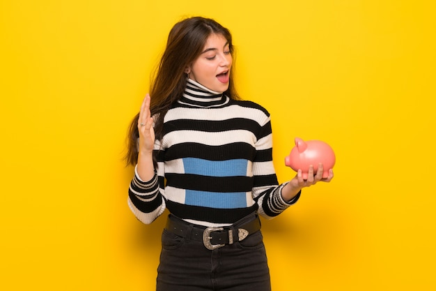 Young woman over yellow wall surprised while holding a piggybank