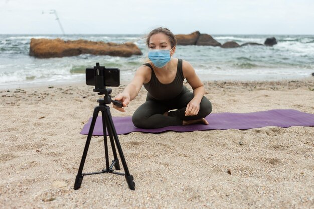Young woman yoga teacher in medical mask leads online lesson at beach Sport woman broadcasts to camera of smartphone standing on tripod Blogging vlog Healthy lifestyle
