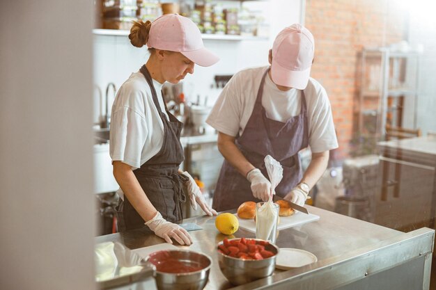 Young womancuts bun with colleague at metal counter in craft bakery shop