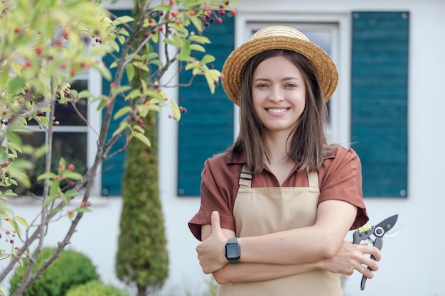 Young womangardener with garden scissors in hand standing in front of garden house