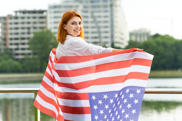 Young womanl holding USA national flag on her shoulders with high office buildings in a city celebrating United States independence day