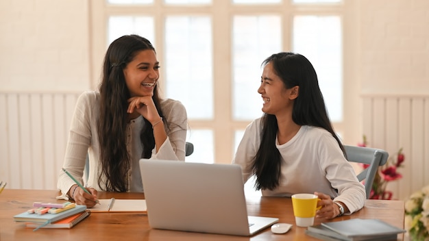 Young women are sitting and smiling at the wooden table surrounded by a computer laptop and equipment