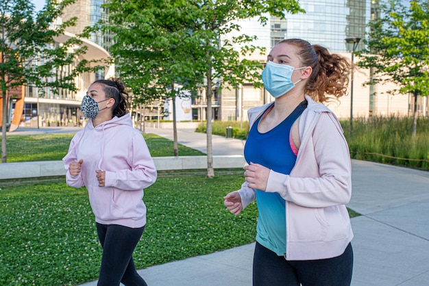 Young women athletes run in the city park, girls exercising in\
the open air wearing protective masks, healthy lifestyle and\
well-being during a coronavirus pandemic