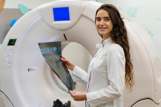 Young women doctor in medical gown looks at results next to CT scanner