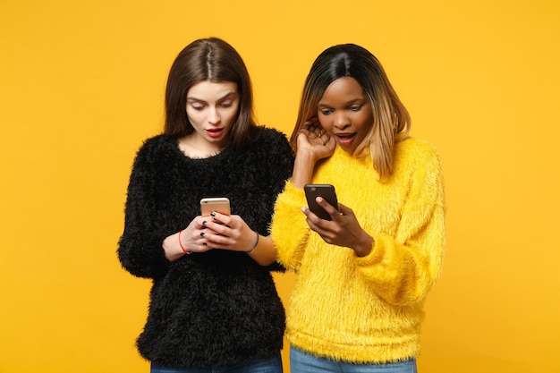 Young women friends european, african american in black yellow clothes hold in hand cellphone isolated on bright orange wall background, studio portrait. people lifestyle concept. mock up copy space
