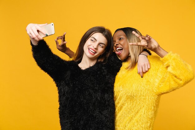 Young women friends european, african american in black yellow clothes hold in hand cellphone isolated on bright orange wall background, studio portrait. People lifestyle concept. Mock up copy space.