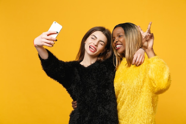 Фото Молодые женщины-друзья европейцы, афроамериканцы в черно-желтой одежде держат в руках мобильный телефон, изолированный на ярко-оранжевом фоне стены, студийный портрет. концепция образа жизни людей. скопируйте пространство для копирования.