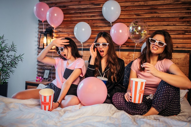 Young women have discgussed emotions on face. They watch movie through special glasses. women sit on bed in festive room. They have pajamas party.