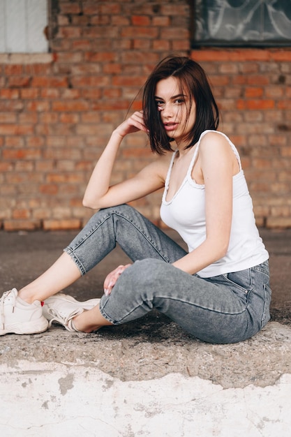 Young women in jeans sneakers sits on concrete in city Model of sexy woman is resting on street