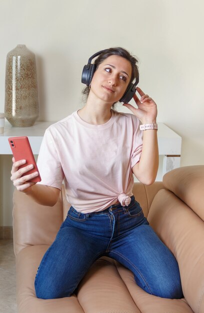 Young women listening to music with headphones and cell phone in hand while sitting on the beige couch at home