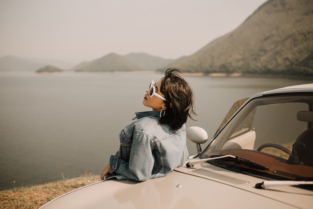 Photo young women relaxing on lake. she go to lake by classic car. she wearing sunglasses.