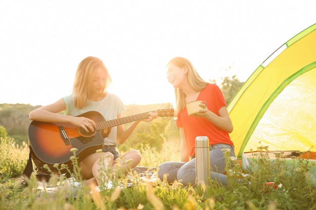 Young women resting with hot drink and guitar near camping tent in wilderness