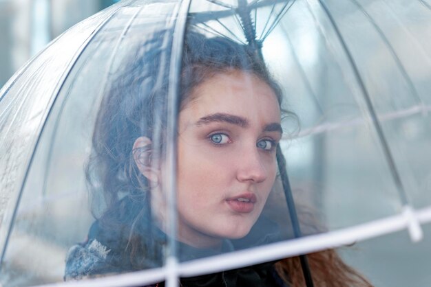 Young women with blue eyes holds transparent umbrella and looks thoughtfully through it at sky Natural beauty