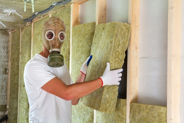 Young worker in gas mask insulating rock wool insulation staff in wooden frame for future house walls for cold and heat barrier. 