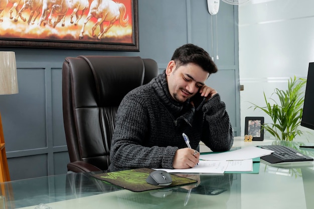 A young worker is sitting in the office doing his work and talking on mobile phone.