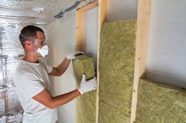 Young worker in respirator insulating rock wool insulation staff in wooden frame for future house walls for cold barrier. Comfortable warm home, economy, construction and renovation concept.