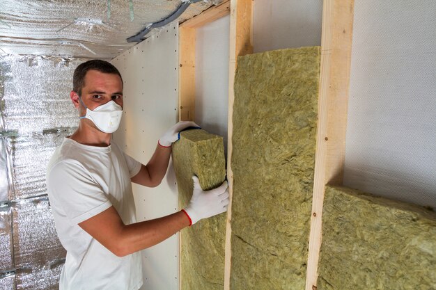 Young worker in respirator insulating rock wool insulation in\
wooden frame walls