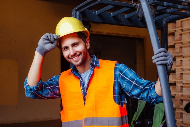 The young worker stands near a forklift in a factory